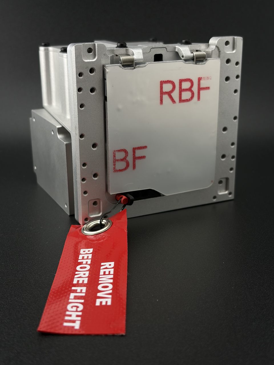 The Astrobrake flight unit, a compact aluminium drag-sail module, with a REMOVE BEFORE FLIGHT tag attached to its release mechanism.