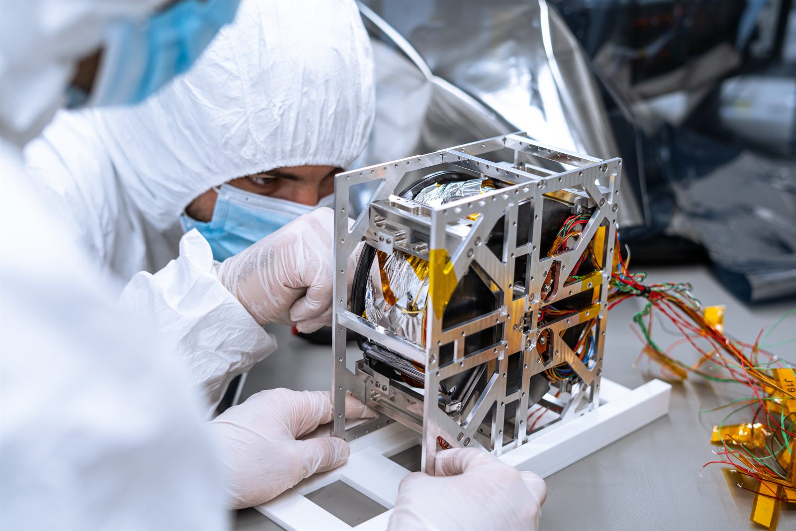 An engineer in cleanroom coveralls, mask and gloves positioning a small satellite structure on an integration jig, its folded membrane and wiring harness visible through the aluminium frame.